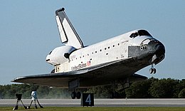 Columbia touches the concrete runway with its rear landing gear at Kennedy Space Center. The tires leave smoke in their wake. Green grass in front of the runway, trees behind, and the blue sky above complement the black and white orbiter.