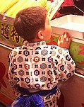 Boy stands at a corn-on-the-cob stall, his back to the viewer; two vertical outward-facing pleats descend from his shoulders.