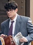 David Corenswet during filming as Clark Kent, wearing glasses and suit and carrying a newspaper and briefcase.