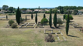 Vestiges au sol, indépendants les uns des autres et de forme rectangulaire, entourés de cyprès et dans un environnement urbain en arrière-plan.