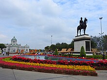 A large plaza with a bronze statue of a man riding on horseback; beyond the plaza is a large two-storey building with a domed roof, arched windows and columns