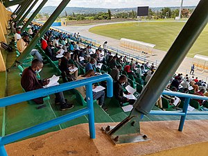 Blick von schräg oben über ein Geländer auf die Ränge des Fußballstadions. Hauptsächlich junge Männer sitzen jeweils mit Abstand auf den Plätzen. Sie tragen Gesichtsmasken und haben Prüfungsbögen auf den Knien, die sie bearbeiten. Im Hintergrund ist noch eine Ecke des Fußballplatzes und die dahinter liegende, locker bewaldete Landschaft zu erkennen.