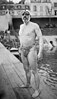 Full body portrait of a man in bathing suit wearing a swim cap and goggles standing on a pier that reaches out into a body of water, with buildings and a few passersby in the background