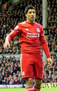 Luis Suárez – wearing a red Liverpool FC jersey with the Standard Chartered sponsor logo at the front centre and shorts with a number 7 partially obscured on the left-leg side and the club crest on the right – lifts his hand with his mouth partly opened.