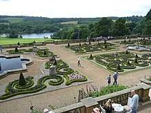 Photo en plongée sur un jardin avec des parterres géométriques et des bassins, avec un lac et des bosquets à l'arrière-plan