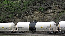 A series of white and black train tank cars with Flammable Hazard placards roll past a steep lush cliffside