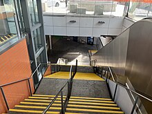 Stairs down to the station concourse viewed from the top. To the left of the stairs is a lift and to the right is an escalator.
