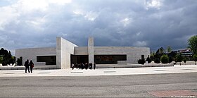 Photographie couleur d’un édifice moderne couleur béton donnant sur un grand parvis sec.