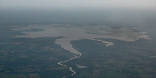 Plan large du lac pris d'avion ; le ciel est brumeux, on distingue les vastes zones de végétation flottante. Au premier plan, un paysage agricole et boisé, avec des zones d'habitation.