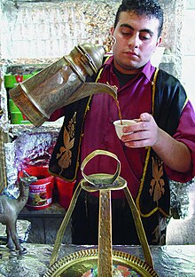 A man with short curly hair, wearing a gold-fringed black vest over a magenta shirt, pours coffee from an ornate, angular container into a small white cup.