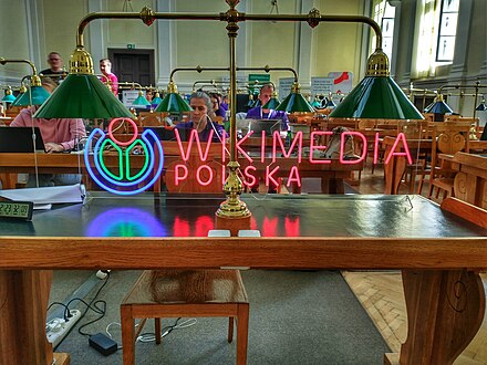 Zdjęcie. Czytelnia w bibliotece, na pierwszym planie neon z napisem "Wikimedia Polska" i lampa. W tle ludzie siedzący przy biurkach i patrzący na laptopy