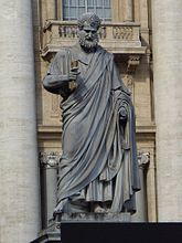 Leichte Untersicht in Farbfotografie von einer Statue, der vor einem alten Gebäude steht. Die männliche Statue trägt eine Stola und hält in ihrer rechten Hand eine Schriftrolle. Er hat lockiges Haar und trägt einen lockigen Vollbart.