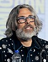 Michael Chabon is pictured wearing glasses, shoulder-length hair and a beard. Speaking at a microphone, his collared shirt is decorated with celestial bodies.