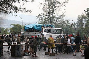 Ein mit UN beschrifteter Lastwagen auf dessen Ladefläche sich unter einer Plane eng gedrängte Menschen befinden, steht auf einer asphaltierten Straße. Er ist von einigen bewaffneten Blauhelmsoldaten umgeben sowie Gruppen von Ruandern, die teils drohend Macheten in Richtung des Lastwagens erheben. Vor und hinter dem Lastwagen sind Absperrungen. Rechts im Bild stehen Mitarbeiter der Filmcrew.