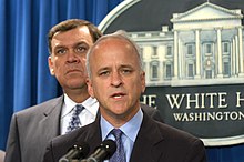 Towey stands at a microphone with a picture of the White House behind him.