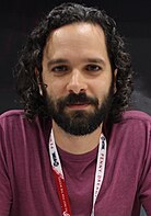 36 year-old man with curly black hair and a beard smirking at the camera.