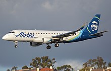 A plane painted with the words 'Alaska' in the front and a blue-green eskimo on the tail soars after take-off just under a cloudy sky