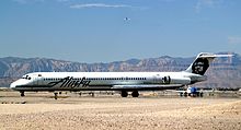 Right side view of an airplane taxiing on the ground towards left side of image. Another plane is behind it, and in the background are mountains and a blue sky with a few clouds.