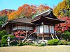 A traditional Japanese building, brown with white shutters, sits behind a field of gravel and in front of multicolored trees