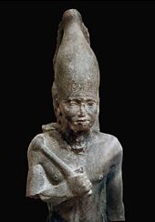 Statue of a standing pharaoh in black stone. He is holding a mace and a rounded crown.