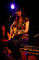 A woman with long black hair wearing a red and black striped singlet. She is seated and playing a guitar in front of a microphone.