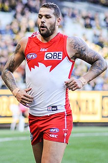 A dark-haired, bearded footballer in a sleeveless red-and-white guernsey running on a grassed oval