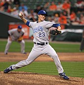 A man in a gray baseball uniform and a blue baseball cap throws a baseball with his right hand. His jersey reads "Kansas City" across the chest in blue script, and "23" underneath in block print. He is wearing a black baseball glove on his left hand, and the blue sleeves of his long undershirt hang down over parts of his forearms.