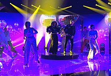 Senhit and Rafał on stage under a disco ball surrounded by backing dancers