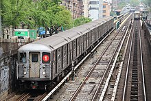 A 1 train, composed here of R62A cars is seen above ground leaving the 125th Street station. The front of the train contains two white lights providing slight illumination, two windows, a door, and the Symbol for the 1 line on the left window.