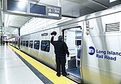 Gleishalle mit aluminiumverschaltem einstöckigem Triebwagen rechts im Bild. Zug mit dem MTA-Logo und dem Text MTA Long Island Rail Road, seitlich Anzeige mit oranger LED-Schrift JAMAICA als Ziel. Anzeigetafel am Bahnsteig hat Aufschrift: Area A / Track 304 / 12:33PM. Mittig im Bild dunkel uniformierter Zugbegleiter, mit Rücken zum Fotografen, seine flache Schirmmütze an einer Hand schwenkend.