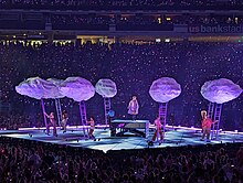 An outdoor stadium filled with spectators on the podiums and on the ground. In the middle Ed Sheeran performs on a raised stage, with video screens above him.