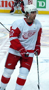 A Caucasian ice hockey player in his thirties. He is skating relaxed on the ice while looking to his right. He wears a white and red jersey, along with a white-visored helmet.