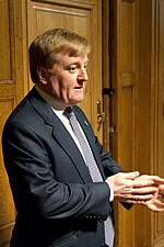 A suited middle-aged man with ginger hair standing in a room with wood paneling. He has a concerned expression and is rubbing his hands.