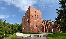 Seitliche Farbfotografie von einem braunen Dom, dessen Mittelteil als Ruine freisteht. In der Bildmitte befindet sich das breite rechteckige Vorderteil mit langen und kurzen Rundbogenfenstern. Der abfallende Putz offenbart hellere Steine. Hinter den rechten Bäumen ist der vordere Bereich des Doms kaum zu sehen.