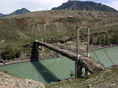 Photographie du pont d'Inya depuis une des deux rives. Le pont se situe dans le fond d'une vallée, même si plus haut d'une dizaine de mètres que la rivière, la Katoun. Le pont est un pont suspendu à haubans, avec deux câbles et deux arches de chaque côté.