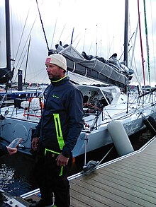 Vêtu d'une combinaison bleu sombre, coiffé d'un bonnet blanc, le navigateur est debout sur le ponton, devant son bateau gris et blanc.