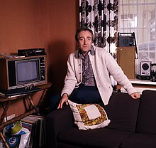 A man in a white cardigan sitting on an armchair posing for the camera with an unexpressed look on his face