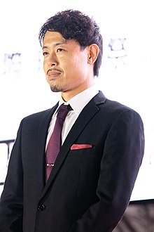 Taguchi at press event in three-quarter profile wearing suit and facing to left of camera