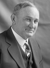 A man with thinning, salt-and-pepper hair wearing a black jacket and vest, white shirt, and patterned tie