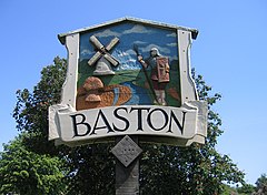 Closeup of village sighn, with a blue sky and green tree in the background. The sign is framed like a medieval barn, around a low relief carving, painted in realistic colours, featuring a stream with a Roman soldier on the right and an 18th century smock mill on the left.