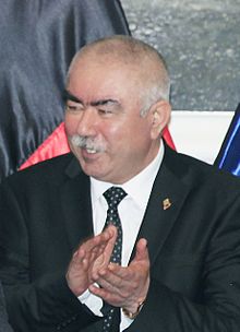Photograph of Abdul Rashid Dostum wearing a black suit with a lapel pin, white shirt, and necktie