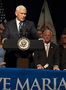 Mike Pence stands at a podium in the foreground; Towey is seated behind him, looking on.