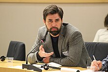 An image of a male delegate seated at a table in a grey jacket with papers and a microphone on the desk.