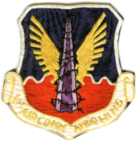 a shield, top half is blue, bottom half red, on it is a grey mololith wrapped in barbed wire with gold eagle wings spread from behind, there is a banner across the bottom with first air commando wing written on it