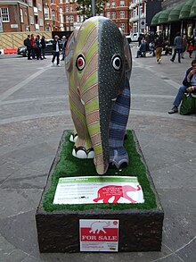 A statue of an elephant is brightly painted in two halves: the left half in lines of yellow and dots of orange; and the right half in overlapping scales in gray, and bands of light and dark blue. The statue sits on a base with the appearance of black rock, an information plate at the front describing the artwork. It is situated in the middle of the pedestrianised road next to Harrods, and people are sitting and walking around in the background.