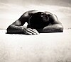 A man lying stomach down on the sand with his head resting on his arm. Taken from a low angle, head-on, in bright sunlight.