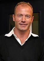 Alan Shearer memakai jumper hitam dengan kolar putih kelihatan.