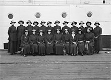 A black and white image of 21 women in dark uniforms of long sleeved jackets, long skirts and hats, posing on the deck of a ship. Nine women are seated in the front row, and twelve are standing behind in the back row. They are standing in front of an outer wall of the ship's cabin, with four portholes spaced out above the standing women.