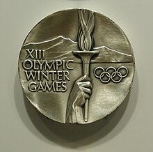 Une médaille avec les mots « XII Olympic Winter Games », une main tenant une torche olympique et des montagnes.