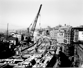 Construction of an elevated highway through an urban neighborhood with mid-rise buildings, seen in black-and-white. A mobile crane is seen towering over workers laying down wood forms.